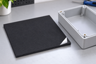 Black silicone sponge square with a white adhesive liner lifting at the corner, grey enclosure box to the right