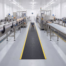 Black 15mm ribbed PVC matting with yellow safety edge ribs laid as a pedestrian walkway between conveyor lines in a food production hall