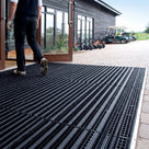 Black brush insert entrance matting, standard grid, recessed flush with the block paving across a golf clubhouse doorway, golfer in muddy shoes stepping onto the bristle strips