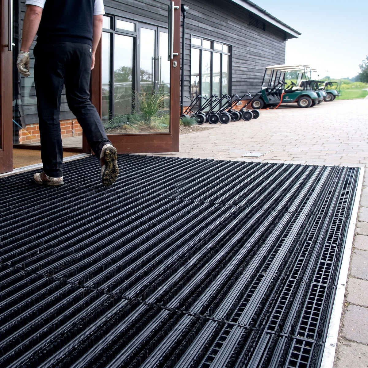 Black brush insert entrance matting, standard grid, recessed flush with the block paving across a golf clubhouse doorway, golfer in muddy shoes stepping onto the bristle strips