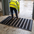 Black brush insert entrance matting, standard grid, recessed flush in a matwell at a warehouse personnel door, worker scrubbing site boots on the bristle strips