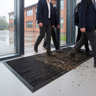 Black brush insert entrance matting, standard grid, recessed across a school entrance on a wet morning with mud scrubbed off pupils' shoes onto the bristles