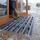 Black brush insert entrance matting, wide grid, recessed flush in a matwell at a farm shop doorway in winter, gravel and slush dropping through the wide open grid