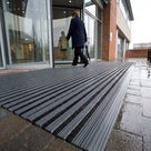 Dark grey brush insert entrance matting, standard grid, outside the revolving door of a hospital main entrance on wet paving