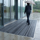 Dark grey brush insert entrance matting, standard grid, recessed flush with the paving at a glazed office entrance in the rain