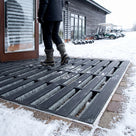 Dark grey brush insert entrance matting, wide grid, recessed flush in a matwell at a snowy clubhouse doorway, snow dropping through the wide open grid