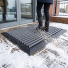 Dark grey brush insert entrance matting, wide grid, at a snowy leisure centre doorway with snow and grit dropping through the wide open grid