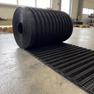 Roll of brush insert entrance matting partly unrolled on a warehouse floor showing the ribbed scraper bars, bristle rows and open grid