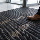 Close-up of black carpet insert entrance matting showing the ribbed PVC scraper bars alternating with absorbent carpet strips, grit in the channels and a shoe stepping on