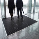 Black carpet insert entrance matting recessed in an aluminium matwell across a glazed office lobby, staff walking in from the rain with wet footprints on the carpet strips