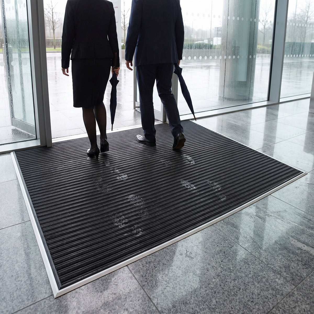 Black carpet insert entrance matting recessed in an aluminium matwell across a glazed office lobby, staff walking in from the rain with wet footprints on the carpet strips