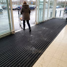 Black carpet insert entrance matting laid as a long run inside a supermarket entrance, shopper pushing a trolley in from a wet car park