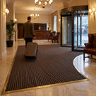 Dark brown carpet insert entrance matting in a brass-edged matwell on a marble hotel lobby floor, porter wheeling a suitcase across it