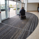 Dark grey carpet insert entrance matting recessed at a hospital entrance, wheelchair user crossing it towards the automatic doors