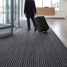 Dark grey carpet insert entrance matting laid as a wide run from a revolving door into a corporate reception, visitor wheeling a suitcase across it