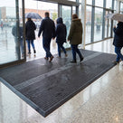 Dark grey carpet insert entrance matting across the full width of a shopping centre entrance, shoppers walking in from the rain