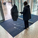 Oxford blue carpet insert entrance matting recessed in a matwell at a library entrance, visitor shaking out a wet umbrella