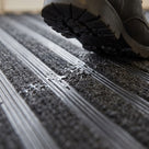 Macro close-up of carpet insert entrance matting: ribbed PVC scraper bars alternating with absorbent carpet strips, rainwater on the pile under a shoe