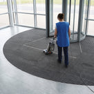 Dark grey carpet insert entrance matting in a revolving door lobby being vacuumed with an upright cleaner