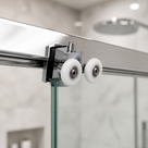 Chrome twin wheel roller clamped to a glass shower panel, its two white wheels running in the enclosure rail
