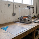 Clear PETG sheet with blue protective film peeled back on a fabrication bench, a curved sneeze guard formed from it beside a strip heater