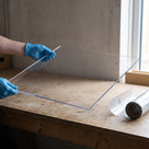 Blue-gloved hands lifting a thin clear polycarbonate sheet above a timber bench, roll of protective film beside it