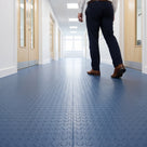 Blue coin-pattern PVC floor matting laid in wide roll lengths down a bright school corridor with oak doors