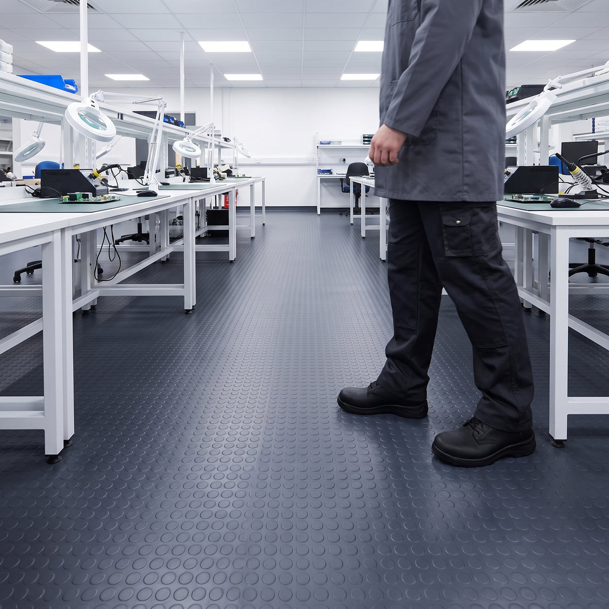 Dark grey coin-pattern PVC floor matting laid wall to wall in a bright, clean electronics assembly room, a technician walking between white benches