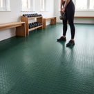 Green coin-pattern PVC floor matting covering a bright boutique fitness studio, a person holding a kettlebell beside oak benches