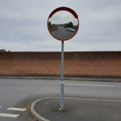 Convex traffic mirror with an orange-red hood on a galvanised pole by a brick wall, reflecting a road and white car