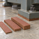 Three loose cork strips of differing thickness on a concrete floor beside a bolted machine foot on a cork pad
