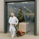 Worker in white coat and hairnet pulls a pallet truck of sacks through a clear PVC strip curtain doorway