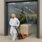 Worker in white coat and hairnet pulls a pallet truck of sacks through a clear PVC strip curtain doorway