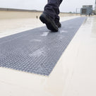 Close view of grey embedded walkway matting flush in a cream liquid roof coating after rain, water draining through the etched open grid under a work boot