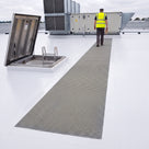 Roof inspector in hi-vis and safety helmet walking a single straight run of grey embedded walkway matting bedded flush into a white liquid-applied roof coating, from the roof hatch to an air-handling unit