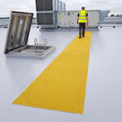 Roof inspector in hi-vis and safety helmet walking a single straight run of yellow embedded walkway matting bedded flush into a pale liquid-applied roof coating, from the roof hatch to an air-handling unit