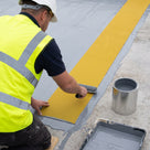 Roofing contractor in hi-vis rolling a strip of yellow open-grid embed walkway matting into wet liquid roof coating so it sits flush with the roof