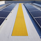Yellow embedded walkway matting set flush into a liquid-coated flat roof as a straight maintenance path between rows of solar panels
