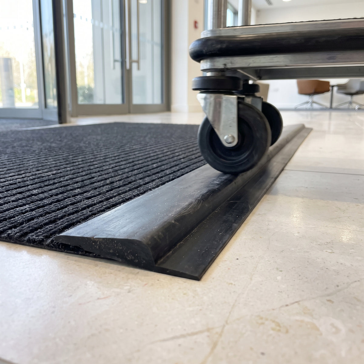 Black PVC ramp edging fitted along the exposed edge of an entrance mat in an office lobby, a trolley wheel rolling up it onto the mat