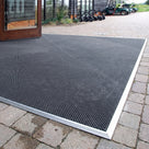 Black fine-ribbed open-grid entrance matting cut to shape with an aluminium edge trim on block paving outside a golf clubhouse