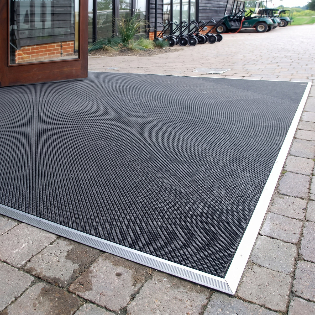 Black fine-ribbed open-grid entrance matting cut to shape with an aluminium edge trim on block paving outside a golf clubhouse