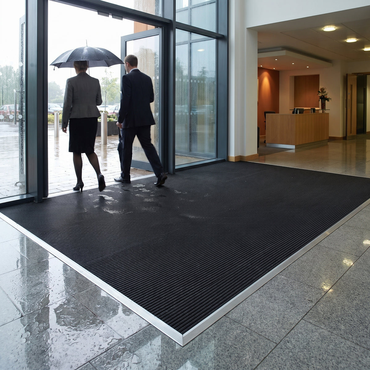 Black fine-ribbed open-grid entrance matting recessed in an aluminium matwell across a corporate office entrance, staff walking in from the rain