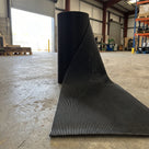 Roll of black fine-ribbed open-grid PVC entrance matting partly unrolled on a warehouse floor, showing the ribbed surface and open grid beneath