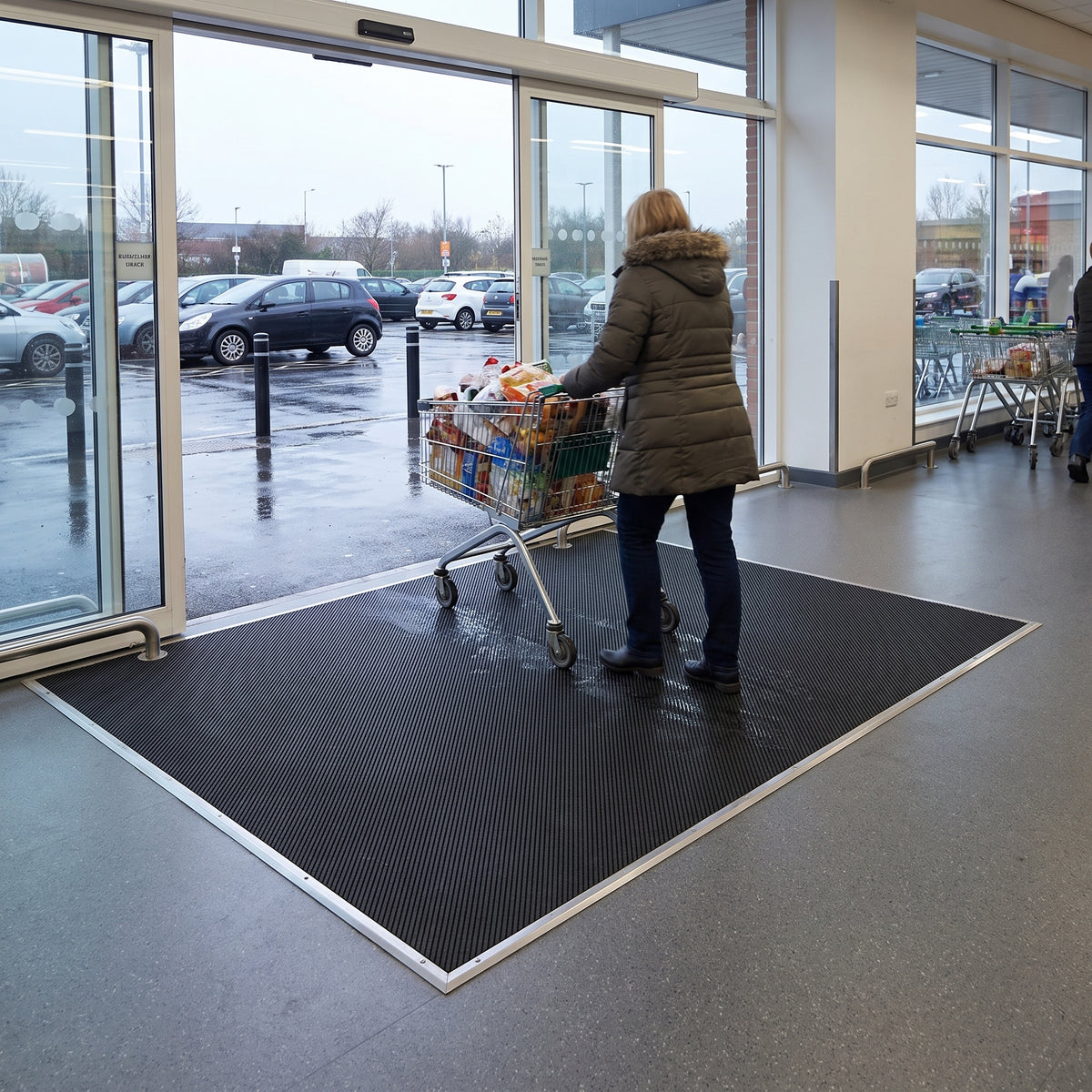 Black fine-ribbed open-grid entrance matting inside a supermarket doorway, shopper pushing a loaded trolley across it from a wet car park