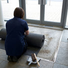 Cleaner rolling back fine-ribbed open-grid entrance matting to sweep the trapped grit out of the matwell underneath