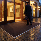 Dark brown fine-ribbed open-grid entrance matting across the timber doorway of a boutique hotel at dusk, porter carrying a bag in
