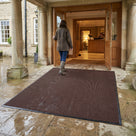 Dark brown fine-ribbed open-grid entrance matting on York stone paving under a country hotel portico, guest stepping in from the rain