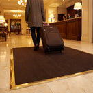 Dark brown fine-ribbed open-grid entrance matting in a brass-edged matwell on a marble hotel lobby floor, guest wheeling a suitcase across it