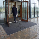 Dark grey fine-ribbed open-grid entrance matting across the timber-framed double doors of an office building, visitor stepping in from wet paving with an umbrella