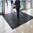 Dark grey fine-ribbed open-grid entrance matting inside the automatic doors of a shopping centre, shopper pushing a trolley across it from the rain